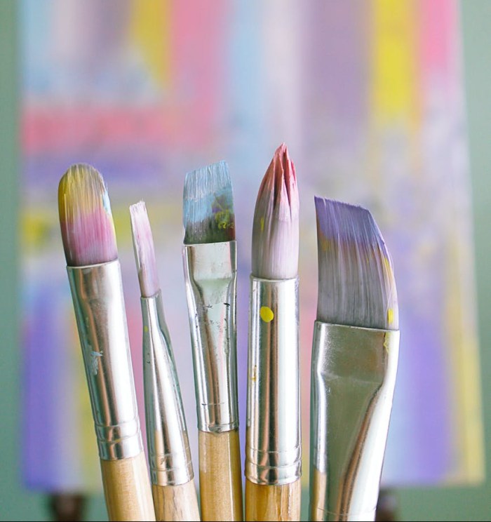 A closeup of paintbrushes which stand vertically, a colorful abstract painting hung behind them