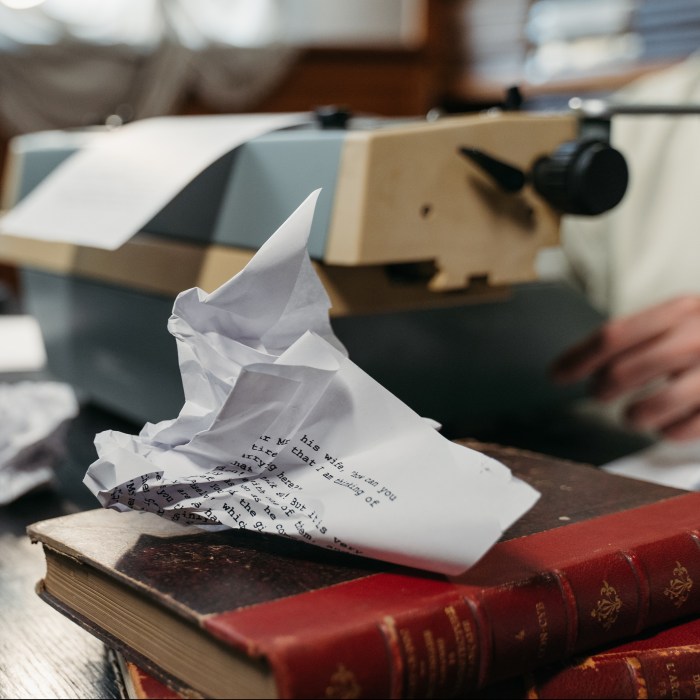 A crumpled piece of paper rests on a stack of books. A typewriter fills the rest of the desk, a hand resting beside it.