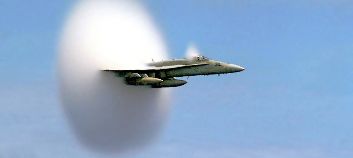 An FA-18 Hornet breaks the sound barrier against a bright, pale sky