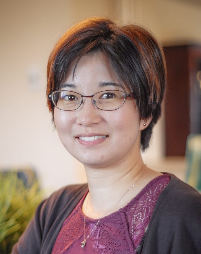 A young Japanese woman with a sweet, round face, glasses, and short-cropped hair stands inside a warm living space, smiling.