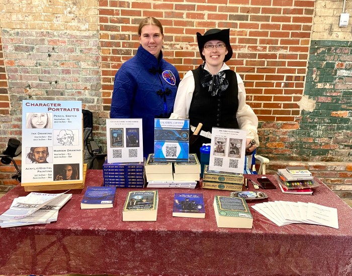 Two young women stand next to each other behind a table displaying books and artwork, cosplaying as fantasy characters.