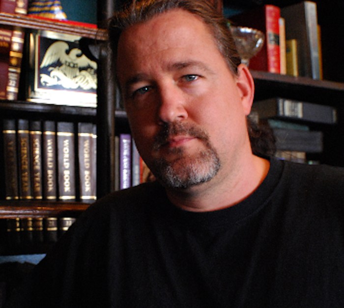 A middle aged man with blue eyes and a peppered beard stands in front of a dark wood bookshelf.