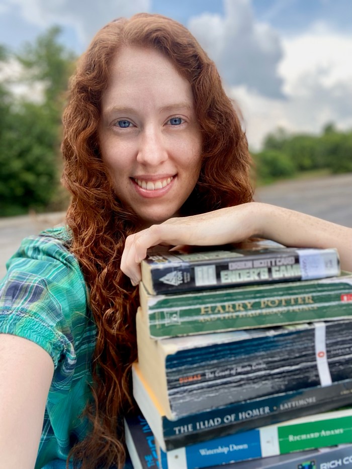 A young woman with curly red hair smiles, her arms resting on a stack of science fiction, fantasy, and classic books